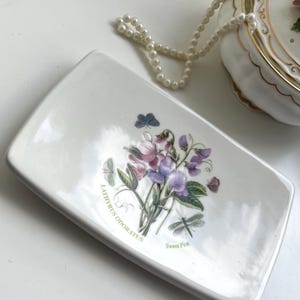Puede incluir: Una jabonera rectangular de cerámica blanca con un diseño floral de guisantes de olor morados y rosas, mariposas y una libélula. El plato tiene el texto "Lathyrus Odoratus" y "Sweet Pea". Un collar de perlas y un recipiente decorativo están en el fondo.