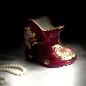 Op de afbeelding: Een diepbordeauxrode, schoenvormige decoratieve keramische houder met een goudkleurige rand en een centrale cartouche met een kleurrijke scène van figuren in historische kleding, versierd met gouden bloemmotieven en een witte binnenkant.