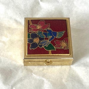 Pequeño anillo cuadrado de metal dorado y esmalte estilo cloisonné para guardar objetos de valor, en una caja vintage.