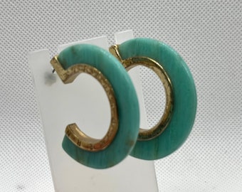 May include: A pair of turquoise and gold hoop earrings. The earrings are made of a light turquoise material with a gold rim. The earrings are about 2 inches in diameter.
