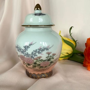 Pode incluir: Um pequeno pote de porcelana branca com tampa. O pote tem um delicado desenho floral em tons de rosa, azul e verde. A tampa tem uma borda dourada e um pequeno desenho de borboleta. O pote está sobre uma superfície branca.