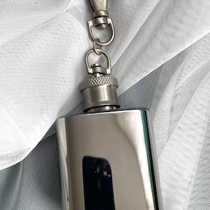 May include: A polished stainless steel hip flask with a silver carabiner clip and chain. The flask is rectangular with a screw-on cap. The background is a white, sheer fabric.