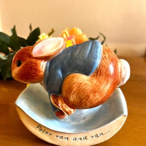 Könnte beinhalten: Eine Keramikfigur von Peter Rabbit aus Beatrix Potters Kinderbuch. Die Figur zeigt Peter Rabbit, der auf einem blauen und weißen Sockel rennt. Der Sockel trägt die Inschrift "Peter ran and ran..."