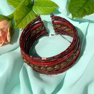 May include: A multi-layered bangle bracelet with a woven brown leather band and red and gold wire accents.