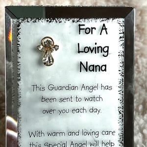 May include: A rectangular glass frame with the text "For A Loving Nana" and a small gold-coloured angel charm. The frame also includes the text "This Guardian Angel has been sent to watch over you each day."