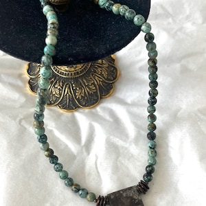 May include: A beaded necklace featuring round, mottled turquoise-coloured stones with dark veining, interspersed with small, dark brown, disc-shaped spacers. The necklace includes a rectangular, flat, dark brown pendant with mottled green accents.