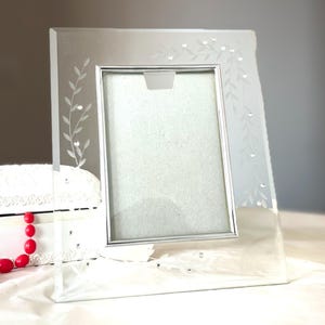 May include: A rectangular glass picture frame featuring a silver-toned inner border and etched leaf patterns along the sides. The clear glass surface is accented with small, embedded clear crystals arranged along the etched vine designs.