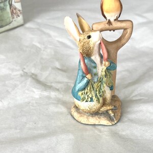 Puede incluir: Una figura de un conejo marrón con una chaqueta azul, sosteniendo un manojo de zanahorias verdes y de pie junto a un mango de herramienta de jardín color canela con un pequeño pájaro marrón posado encima, sobre una base circular clara.