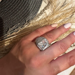 Puede incluir: Un anillo de plata con una piedra grande, cuadrada y transparente rodeada de piedras más pequeñas. El anillo está en el dedo de una persona.