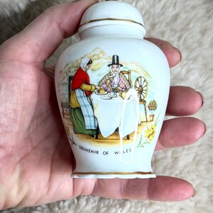Può includere: Vaso in ceramica bianca con coperchio dorato e una scena dipinta di due persone sedute a un tavolo con una ruota da filatura sullo sfondo. Il vaso ha la scritta "Souvenir of Wales" sul fondo.