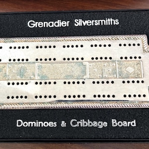 Puede incluir: Un juego de dominó y cribbage plateado en una caja negra. El tablero tiene numerosos agujeros para clavijas y un borde decorativo. La caja está etiquetada como "Grenadier Silversmiths" y "Dominoes & Cribbage Board".