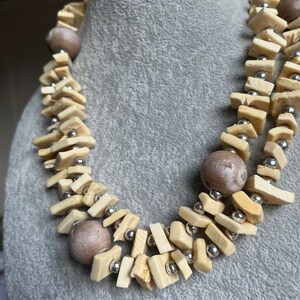 May include: A multi-strand necklace made of light brown and cream colored wooden beads and silver metal beads. The necklace is layered and features a variety of shapes and sizes of beads.