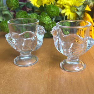 May include: Two clear glass egg cups, each shaped like a bird. The cups have a round bowl for the egg and are supported by a bird-shaped base with a detailed wing and tail design. The background features yellow and green flowers.