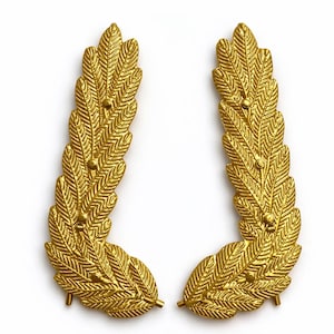 Könnte beinhalten: Zwei goldfarbene, blattförmige Dekorationselemente. Jedes Element hat eine detaillierte, strukturierte Oberfläche mit einer Reihe kleiner, runder Akzente. Das Design ist symmetrisch, mit einer geschwungenen Form, die an einen Lorbeerkranz erinnert. Der Hintergrund ist weiß.