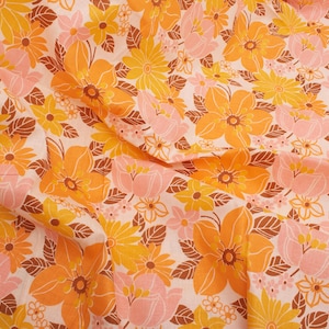 May include: Close-up of a fabric with a repeating floral pattern. The design features large orange and yellow flowers, smaller pink and yellow blossoms, and brown leaves on a white background. The fabric is folded.