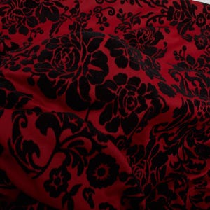 May include: Close-up of a red fabric with a black floral pattern. The design features large, stylized roses and other floral elements. The fabric has a soft, velvety texture and a rich colour palette.