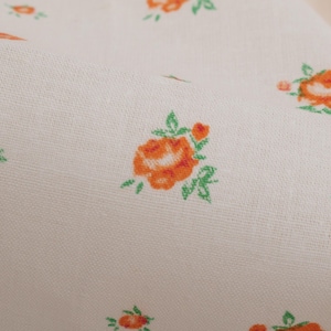 以下が含まれることがあります： クリーム色の生地のクローズアップ。オレンジ色のバラと緑の葉の繰り返し模様があります。生地はわずかにテクスチャ加工されており、バラのサイズと詳細が異なります。