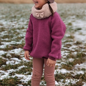 Könnte beinhalten: Ein junges Mädchen trägt eine rosa Strickmütze mit Bommel, einen rosa Strickschal, einen rosa Pullover, rosa Leggings und rosa Stiefel. Sie steht auf einem schneebedeckten Feld.