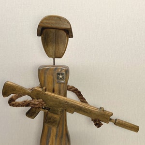 May include: Wooden figurine of a soldier wearing a brown hat and holding a wooden rifle. The soldier is wearing a brown uniform with a U.S. Army patch.