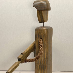 May include: A wooden figurine of a soldier with a rifle and a rope. The soldier is wearing a hat and has a brown and gray wood grain finish.