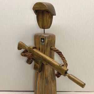 May include: Wooden figurine of a soldier wearing a brown hat and holding a brown rifle. The soldier is wearing a U.S. Army patch on the chest.