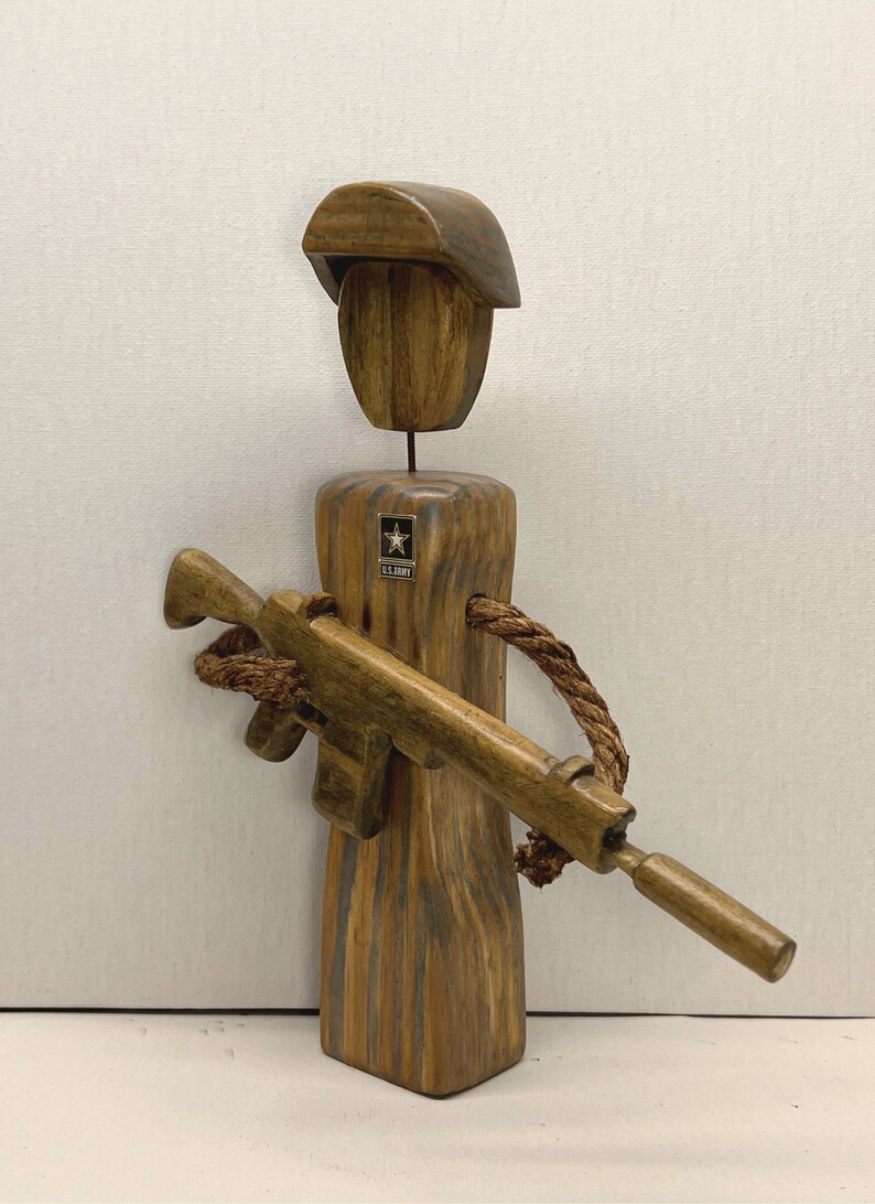 May include: Wooden figurine of a soldier wearing a brown hat and holding a brown rifle. The soldier is wearing a U.S. Army patch on the chest.