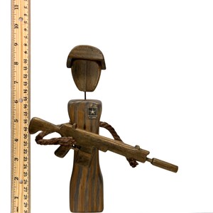 May include: A wooden figurine of a soldier wearing a helmet and holding a rifle. The soldier is wearing a U.S. Army patch on the chest. The figurine is painted in shades of brown and black.