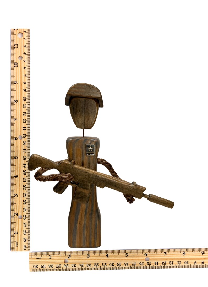 May include: A wooden figurine of a soldier wearing a helmet and holding a rifle. The soldier is wearing a U.S. Army patch on the chest. The figurine is painted in shades of brown and black.