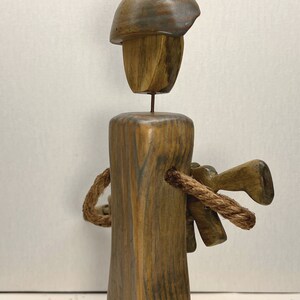 May include: Wooden sculpture of a person wearing a helmet and holding a rope. The sculpture is made of brown wood with a dark wood grain. The person is standing on a wooden base.