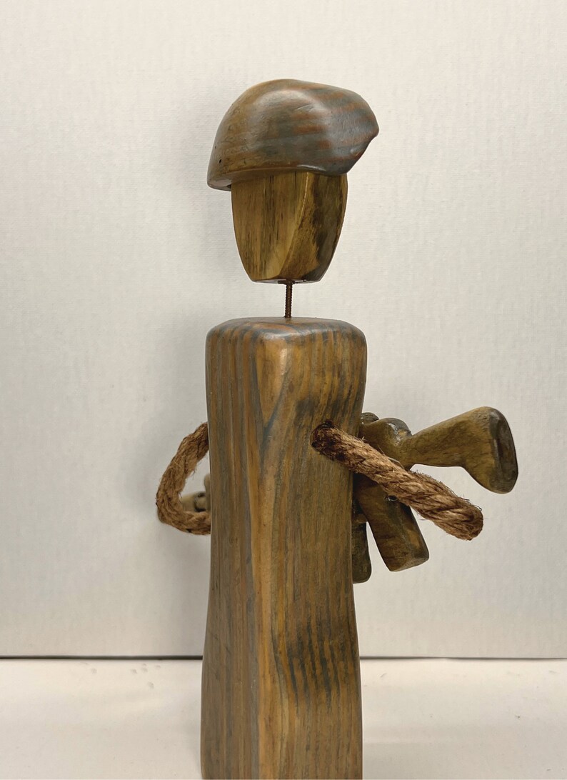 May include: Wooden sculpture of a person wearing a helmet and holding a rope. The sculpture is made of brown wood with a dark wood grain. The person is standing on a wooden base.