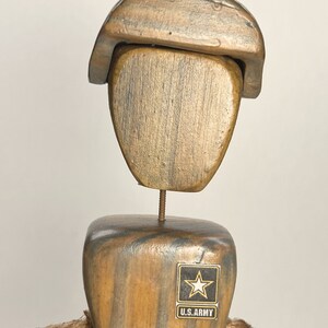 May include: Wooden figurine of a soldier wearing a brown hat and a U.S. Army patch. The soldier is holding a wooden rifle and is standing on a wooden base.