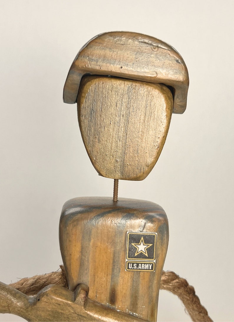 May include: Wooden figurine of a soldier wearing a brown hat and a U.S. Army patch. The soldier is holding a wooden rifle and is standing on a wooden base.