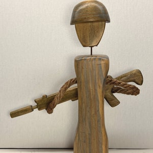 May include: A wooden figurine of a soldier with a helmet, a rifle, and a rope tied around the rifle. The figurine is made of wood with a brown and gray finish.