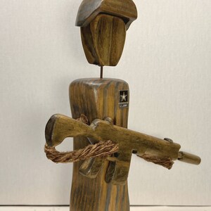 May include: Wooden figurine of a soldier wearing a helmet and holding a rifle. The soldier is wearing a U.S. Army patch on the chest.
