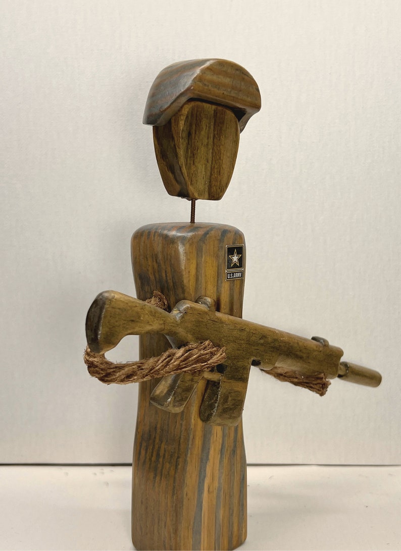 May include: Wooden figurine of a soldier wearing a helmet and holding a rifle. The soldier is wearing a U.S. Army patch on the chest.