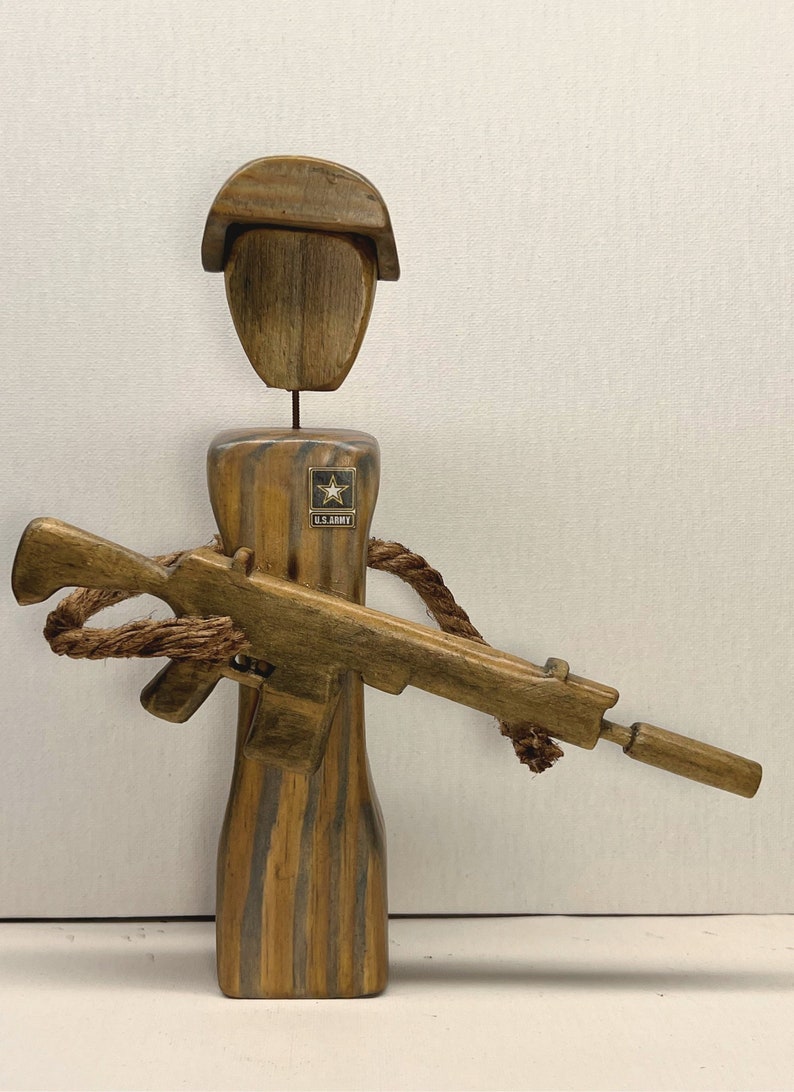 May include: Wooden figurine of a soldier wearing a brown hat and holding a wooden rifle. The soldier is wearing a brown uniform with a U.S. Army patch.
