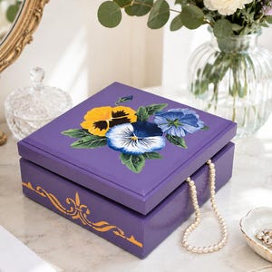 May include: A square purple wooden box featuring a painted cluster of yellow, white, and blue pansies on the lid. The front side of the box is decorated with a gold-coloured filigree design. A string of white pearls hangs from the open lid.