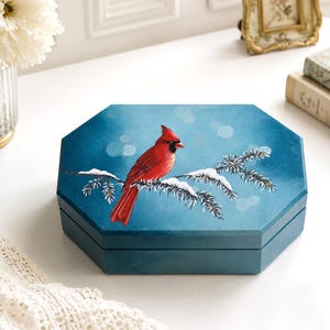 May include: An octagonal wooden box painted in a deep blue hue, featuring a detailed illustration of a bright red cardinal bird perched on a snow-covered pine branch against a soft, bokeh-effect blue background on its lid.