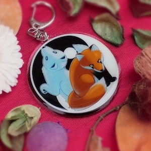 Peut inclure: Un porte-clés rond et transparent avec un symbole yin yang noir et blanc. Un ours polaire blanc et un renard orange sont représentés sur le symbole. L'ours polaire est sur le côté blanc du yin yang et le renard est sur le côté noir.
