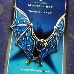 Könnte beinhalten: Ein Hard-Emaille-Pin mit einem mystischen Fledermaus-Design mit dunkelblauen Glitzerflügeln und goldenen Akzenten. Der Pin befindet sich auf einer blauen Karte mit dem Text "AZILORD MYSTICAL BAT IN DARK GLITTER".