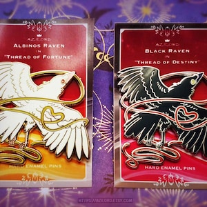 Peut inclure: Deux épingles en émail dur représentant des corbeaux. L'une est blanche avec des détails dorés, l'autre est noire avec des détails dorés. Les deux épingles sont sur des cartes rouges avec le texte "Albino's Raven in Thread of Fortune" et "Black Raven in Thread of Destiny".