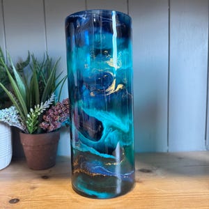 May include: A cylindrical glass vase featuring a swirling marbled pattern in shades of deep navy blue, bright turquoise, and teal, accented with metallic gold streaks throughout the surface. The vase has a smooth, glossy finish and a straight shape.