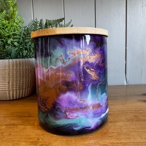May include: A cylindrical jar with a wooden lid. The jar's exterior features a swirling design in shades of purple, teal, and gold. The jar is sitting on a wooden surface.