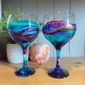 Puede incluir: Dos copas de vino transparentes con diseños abstractos vibrantes. Los interiores presentan patrones arremolinados de color turquesa, morado y dorado. Los tallos y las bases hacen la transición del azul al morado. Las copas están sobre una superficie de madera.