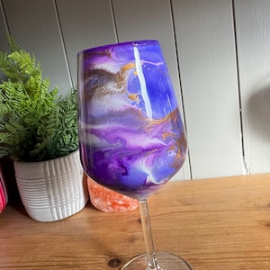 May include: A wine glass with a stem, featuring a vibrant, abstract design. The glass is adorned with swirling shades of purple, blue, and gold, creating a unique, artistic aesthetic. The glass is clear, allowing the colors to shine through.
