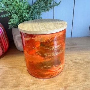 Puede incluir: Un tarro cilíndrico de vidrio con tapa de bambú. El exterior del tarro presenta un diseño veteado naranja y dorado. La tapa es de color beige claro. El tarro está sobre una superficie de madera.