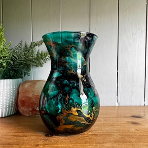 May include: A glass vase featuring a swirling marbled pattern of deep emerald green, black, and metallic gold accents. The vessel has a wide, rounded body that tapers towards a slightly flared rim, showcasing a smooth, glossy finish throughout.