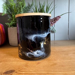 Może przedstawiać: Czarny ceramiczny pojemnik z bambusową pokrywką. Pojemnik ma błyszczące wykończenie z białym i złotym marmurkiem. Pojemnik jest cylindryczny i zawiera zieloną roślinę. Pojemnik stoi na drewnianej powierzchni.