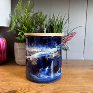 May include: A cylindrical jar with a wooden lid, showcasing a swirling design of deep blue, white, and gold resin. The jar is approximately 15 cm tall and 10 cm in diameter, ideal for storage or decorative purposes.