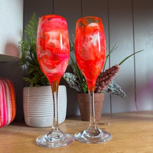 Puede incluir: Dos copas de champán con diseños abstractos vibrantes en rojo, rosa y blanco. Los tallos son transparentes y las bases redondas. Las copas se exhiben sobre una superficie de madera, sugiriendo un tema festivo o decorativo.